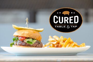 A Symphony of Lights-themed burger and fries on a plate with the words cured table & tap.